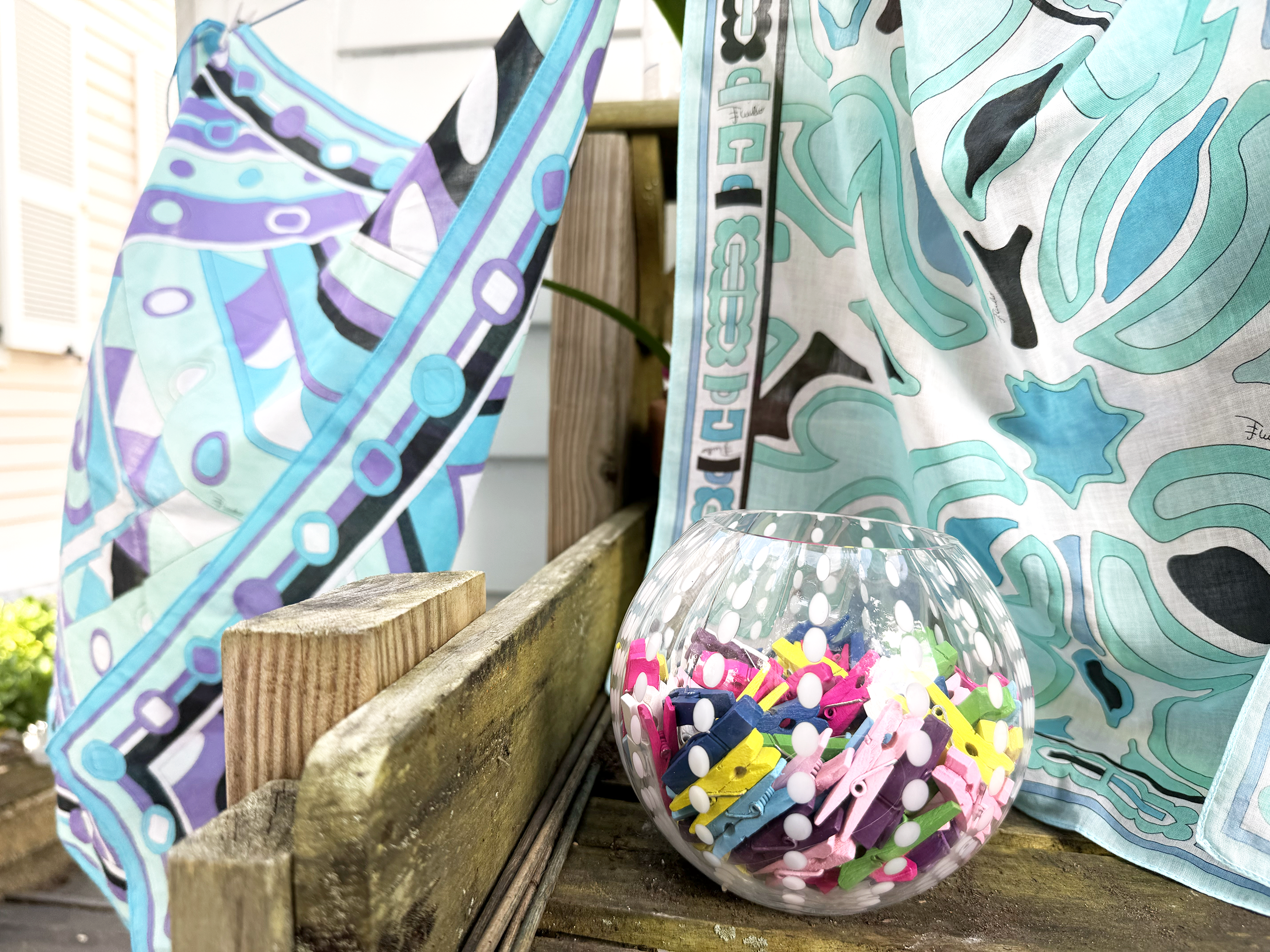 A small glass bowl of miniature clothespins in front of scarves on the clothesline.