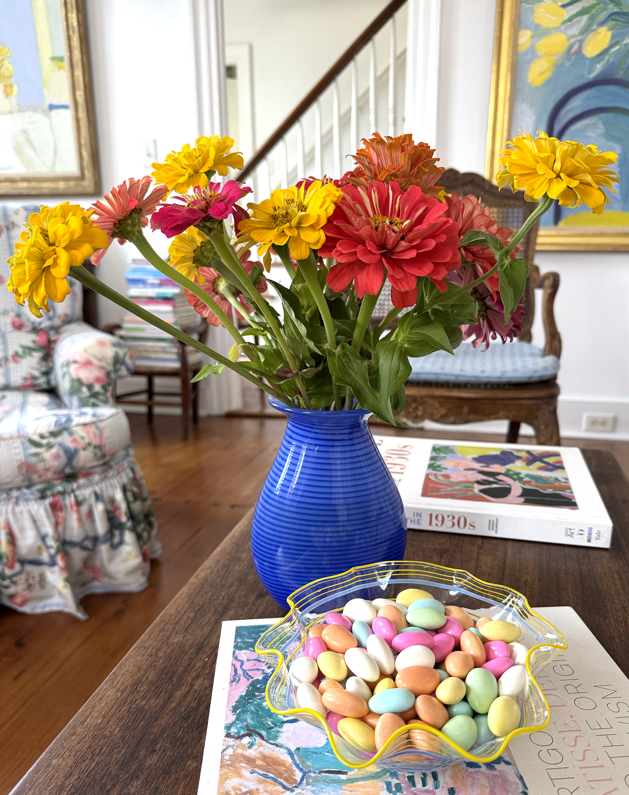 A dish of Jordan almonds on top of an art book next to a vase of colorful dahlias.
